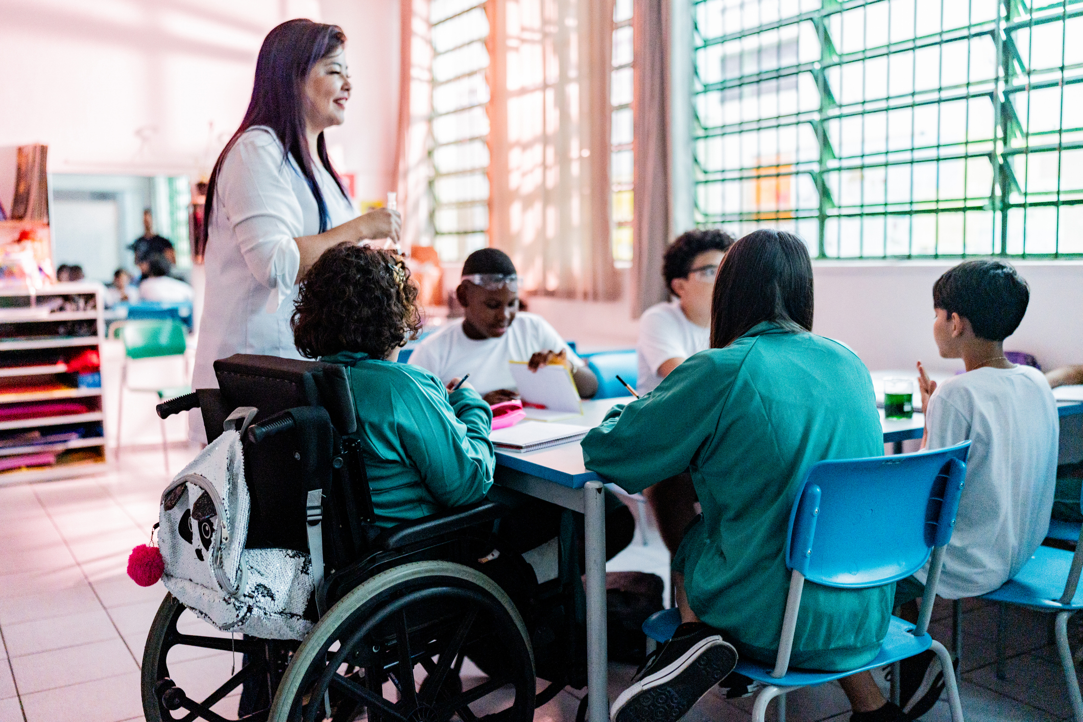 A teacher talks to five students at a table, one of whom is sitting in a wheelchair.