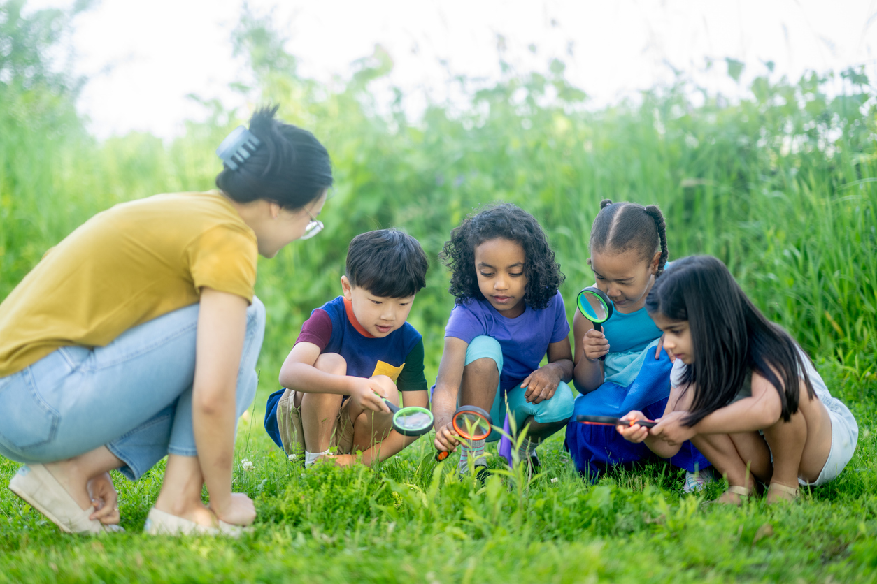 A teacher kneels next to four children looking through magnifying glasses in a grassy field. 